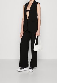 Schwarzer ärmelloser Blazer und weit geschnittene Hose mit einem tiefen Ausschnitt, kombiniert mit einer weißen Handtasche und schwarzen Sneakers. Glatte Stofftextur.