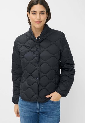 Woman wearing a black quilted jacket with snap buttons and blue jeans, standing with one hand in pocket against a plain background.