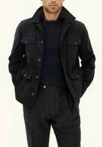 Giacca in cotone blu navy con colletto alto, quattro tasche frontali e chiusura a bottoni, abbinata a un maglione scuro con texture e pantaloni neri.
