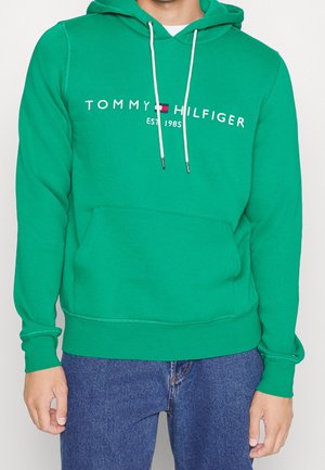 Grüner Tommy Hilfiger Hoodie mit weißen Kordeln, Logo und dem Text "EST. 1985", getragen mit blauen Jeans, Vorderansicht.