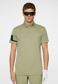 Olivgrön pikétröja med korta ärmar, krage och knapplist med tre knappar; har en svart logotyp på vänster ärm och en jämn textur.