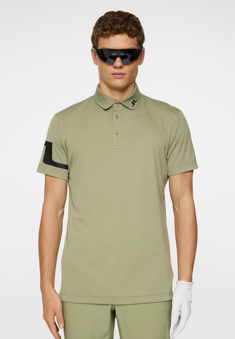 Olivgrön pikétröja med korta ärmar, krage och knapplist med tre knappar; har en svart logotyp på vänster ärm och en jämn textur.