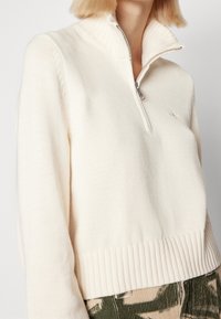 Kvinna som bär en krämfärgad stickad tröja med halvdragkedja och ribbad nederkant, i kombination med gröna och beige mönstrade byxor.