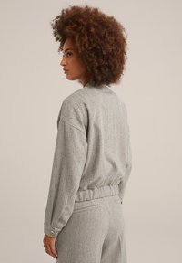 Costum texturat gri cu o croială lejeră, prezentând un guler, umeri căzuți și talie elastică. Model herringbone notabil și mansete cu nasturi.