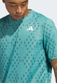 Lett teal treningstopp med et geometrisk mønster av mørke og lyse diamanter, rund halslinning og hvit logo.