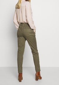 Olijfgroene tapered broek met een hoge taille, achterzakken en een zachte katoenmix stof, gecombineerd met een licht gestreept blouse en bruine haklaarzen.