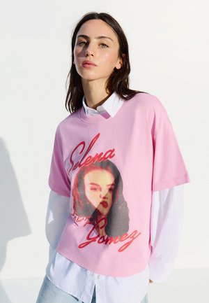 Junge Frau trägt ein übereinander getragenes weißes Hemd und ein rosa Selena Gomez Grafik-T-Shirt, steht vor einem weißen Hintergrund mit Schatten.