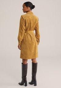 Rochie cămașă din corduroy galben muștar, cu mâneci lungi, talie încinsă și manșete cu nasturi; asortată cu cizme de piele negre până la genunchi.