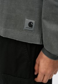 Szary bawełnowy sweatshirt z ribbowanym mankietem, z wykończonym szwami dołem oraz prostokątną, szaro-czarną łatą z logo.