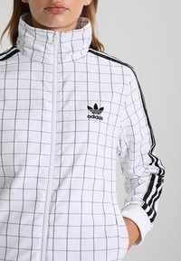 Kvinna i en vit Adidas zip-up jacka med svart rutmönster och svarta ränder längs ärmarna, med handen i fickan mot en grå bakgrund.