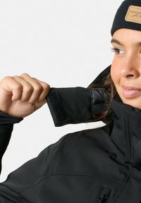 Zwarte jack met een hoge kraag, met een klittenbandriem op de pols. Het materiaal lijkt soepel en duurzaam, met een ritsluiting.