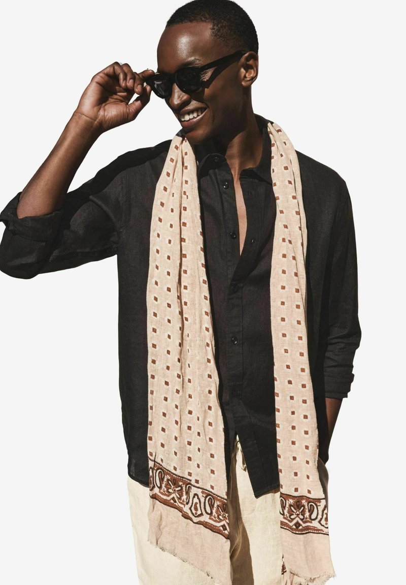 Personne souriante portant des lunettes de soleil foncées, une chemise noire boutonnée, une écharpe claire à motifs et un pantalon clair, tenant les lunettes de soleil d'une main.