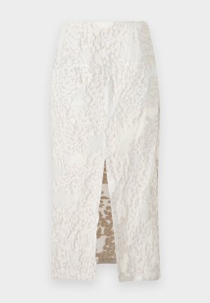 Vrouw draagt een gestructureerde witte lange mouwen jas en bijpassende midi rok met voorste split, gecombineerd met zwarte open teen hakken met gouden accenten.