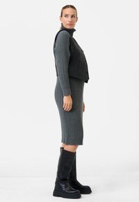 Graues geripptes Strickkleid mit hohem Kragen, darunter eine schwarze, kurz geschnittene Lederweste, kombiniert mit hohen schwarzen Stiefeln mit dicken Sohlen.