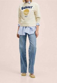 Sudadera crema con el texto "Sunset" y un gráfico amarillo, superpuesta sobre una camisa de rayas azul claro, combinada con unos vaqueros acampanados azul claro y tacones verdes.