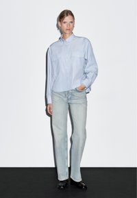 Camicia azzurra chiara a righe con due taschini sul petto, abbinata a jeans a gamba dritta di lavaggio chiaro e scarpe nere su uno sfondo scuro.