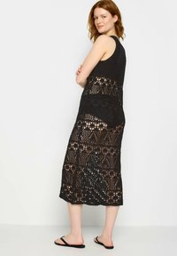 Vestido midi de ganchillo negro que presenta un diseño sin mangas con un patrón calado. La prenda tiene un corte holgado y un acabado texturizado.