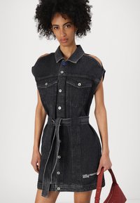 Μαύρο τζιν φόρεμα με κοντά μανίκια, κούμπωμα με κουμπιά μπροστά, ζώνη στη μέση, δύο τσέπες στο στήθος και λεπτομέρεια λογότυπου. Διαθέτει λευκό ραφές σε αντίθεση.