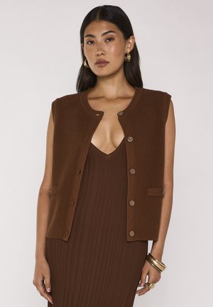 Donna con capelli lunghi e scuri indossa un gilet marrone in maglia senza maniche con bottoni sopra un vestito marrone a coste abbinato e orecchini a cerchio e braccialetti dorati.