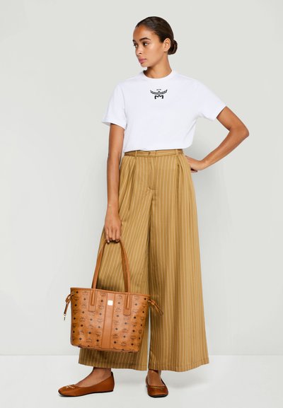 Femme portant un t-shirt avec logo blanc et un pantalon large à rayures marron, tenant un sac fourre-tout à motifs marron, avec des ballerines marron assorties.