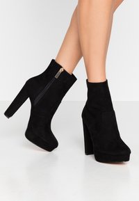 Czarne zamszowe botki za kostkę z grubym wysokim obcasem, zamkiem błyskawicznym po boku i gładką teksturą. Projekt platformy zwiększa wysokość.