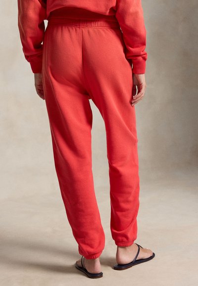 Osoba ubrana w czerwone spodnie dresowe i pasującą bluzę, stojąca boso w czarnych sandałach na neutralnym tle.