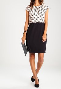 Eine Frau trägt eine Bluse mit Punkten und einen schwarzen Rock, hält eine schwarze Clutch und trägt schwarze High Heels, während sie vor einem weißen Hintergrund steht.