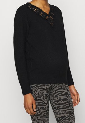 Person iført en sort sweater med blonder V-halsdetalje og grå mønstrede bukser med sorte abstrakte striber.