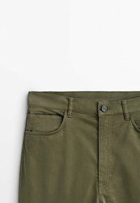 Pantalon vert olive avec fermeture à boutons sur le devant, passants de ceinture et deux poches avant sur fond blanc uni.