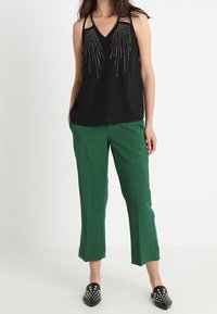 Black sleeveless top with beaded fringe detail, paired with green, cropped trousers and black slip-on shoes with studs.
