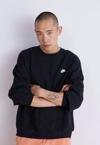 Fekete Nike pulóver, kerek nyakkal, ejtett vállakkal és bordázott mandzsettákkal. A mellkason egy kis fehér logó található. Simább textúra, laza fazon.