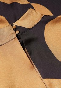 Une blouse en soie présentant un motif abstrait noir et or, ornée de deux boutons ronds au col et d'une texture de tissu lisse et fluide.