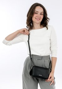 Schwarze Leder-Crossbody-Tasche mit strukturierter Oberfläche, verstellbarem Tragegurt und silbernen Beschlägen, getragen zu einem weißen Oberteil und grauen Hosen.