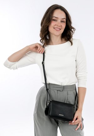 Černá kožená crossbody taška s texturovaným povrchem, nastavitelným popruhem a stříbrným kováním, nošená s bílým topem a šedými kalhotami.