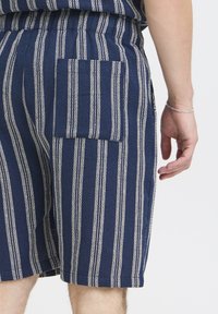Tmavomodré krátke nohavice s bielymi zvislými pruhmi, s náprsným vreckom na ľavej strane a textúrovanou tkaninou. Zobrazený elastický pás.
