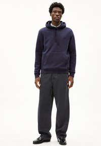 Haut à capuche marine avec une poche kangourou et des cordons, associé à un jean gris foncé ample. Le mannequin se tient sur un fond blanc uni.