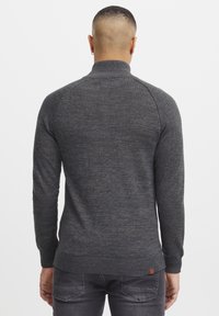 Maglione grigio scuro lavorato a maglia con colletto alto, vita e polsini a costine. Presenta un tessuto texturizzato e un piccolo etichetta con logo vicino all'orlo.