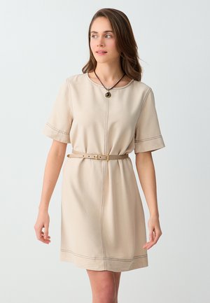 Kobieta o jasnobrązowych włosach, ubrana w beżową sukienkę z krótkim rękawem i paskiem oraz naszyjnik z wisiorkiem, stojąca na jednolitym białym tle.