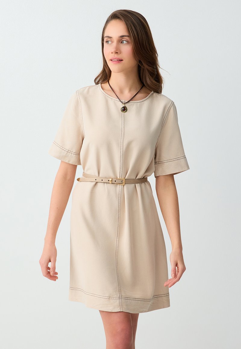 Femme aux cheveux châtain clair portant une robe beige à manches courtes ceinturée et un collier avec pendentif, debout devant un fond blanc uni.