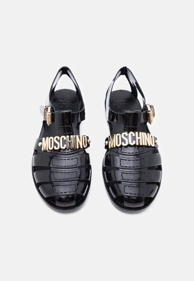 Sandales en gelée noires brillantes avec logo Moschino doré sur les brides avant et boucles dorées, vues de dessus sur fond blanc.