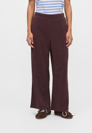 Personne portant un pantalon ample violet foncé, des chaussures marron à lacets et un haut rayé bleu et blanc, debout devant un fond uni.