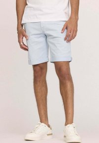 Shorts en denim bleu clair avec des ourlets retroussés, présentant un design classique. Portés avec des baskets blanches, sur un fond uni.