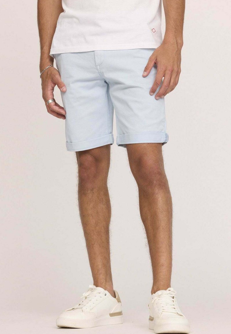 Shorts en denim bleu clair avec des ourlets retroussés, présentant un design classique. Portés avec des baskets blanches, sur un fond uni.