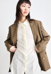 Bruine, op maat gemaakte blazer over een witte, knoopshirt. De blazer heeft schoudervullingen en een grote gele knoop. Soepele, lichte stof.