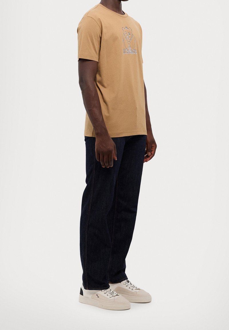 Homme portant un t-shirt beige avec un motif d'ours, un jean bleu foncé et des baskets blanches, debout devant un fond blanc uni.