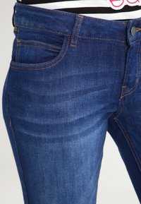 Tmavě modré džíny z denimu s úzkým střihem, ozdobené oranžovým prošitím, s přední kapsou a hladkým povrchem. Kovové zapínání na knoflík v pase.