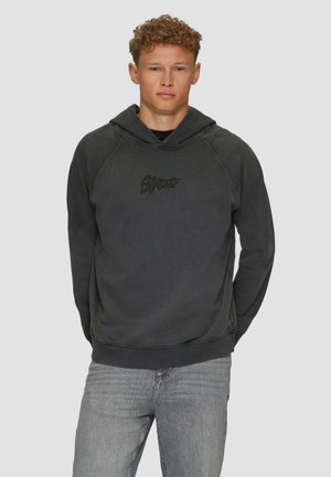 Zwarte hoodie met een kangoeroezak, gestructureerde stof en een logo aan de voorkant. Beschikt over een verstelbare capuchon en geribde manchetten. Gecombineerd met grijze jeans.