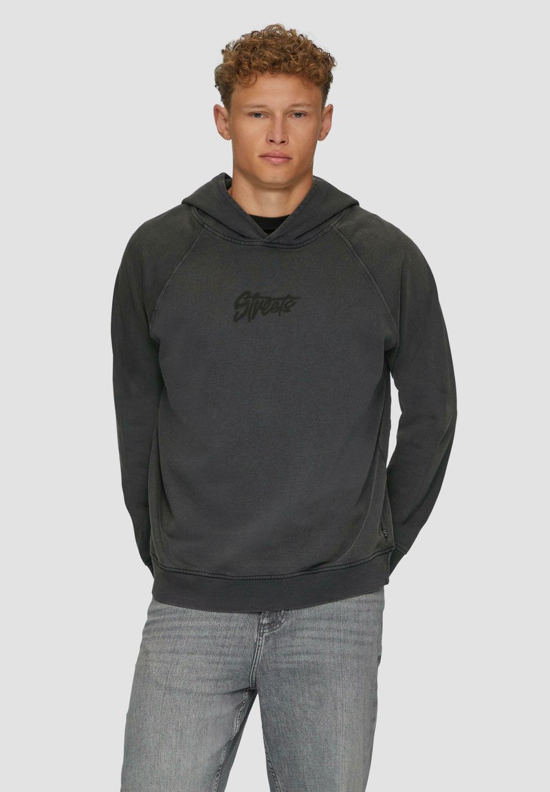 Czarny hoodie z kieszenią kangurką, teksturowana tkanina i logo z przodu. Posiada sznurki w kapturze oraz ściągacze na mankietach. Noszony z szarymi dżinsami.