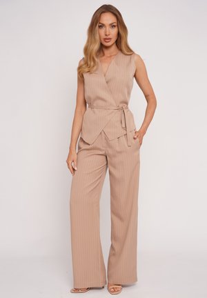 Frau mit langen blonden Haaren, die eine beige, ärmellose Wickeltop mit Nadelstreifen und passende weit geschnittene Hosen trägt, steht vor einem schlichten Hintergrund.