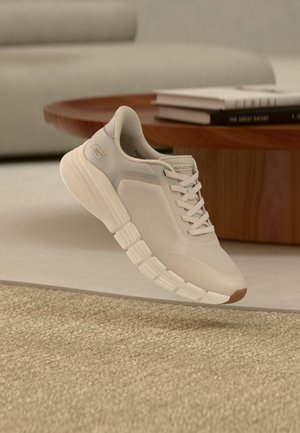 Lys beige Skechers-sneaker med tyk, opdelt sål og meshpaneler, placeret nær et træbord med stablede bøger.